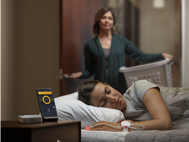 Concerned mother holding laundry basket in bedroom doorway while watching over daughter wearing Opioid Halo sensor in bed, with smartphone and Home Medical Hub at bedside 