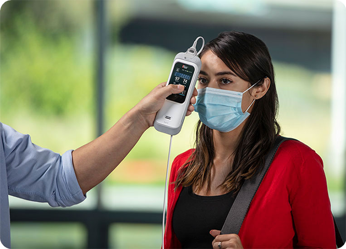 A healthcare provider checks a masked woman’s temperature using a Masimo Rad-G with Temperature device in an indoor setting.